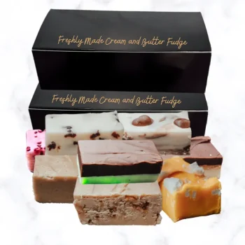 Two black boxes of fudge with bars of fudge stacked in front of the boxes, all displayed on a white marble background